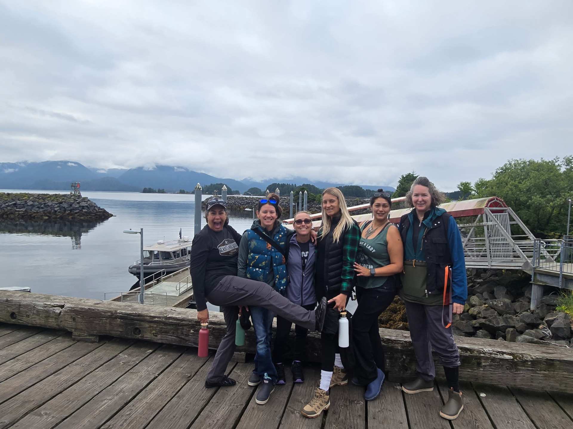 Six women posing on a wooden dock in front of the Sitka marina